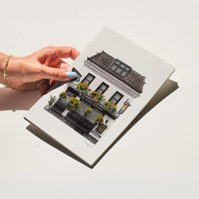 hand with blue nail polish holding a print featuring an illustration of The Mayflower pub with hanging flower baskets against a white background.