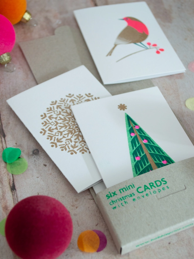 A boxed set of six festive Christmas cards lies open and three mini Christmas cards printed with Christmas Trees, Snowflakes and festive Robins are spilling out onto a wooden table. Small brightly coloured baubles are scattered around the box and Christmas cards.