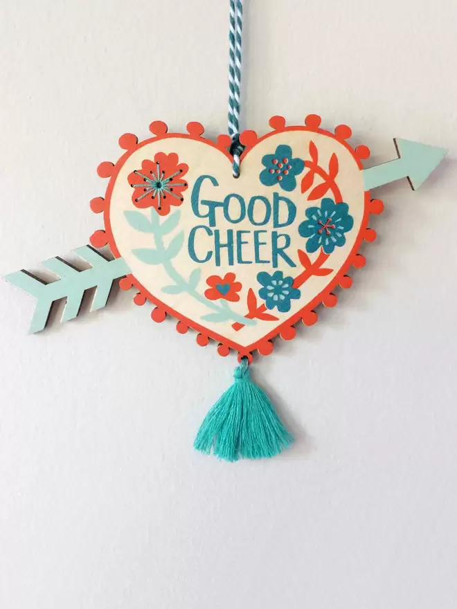 'Good Cheer' is hand printed on a blue and red floral wooden heart with an arrow through it.