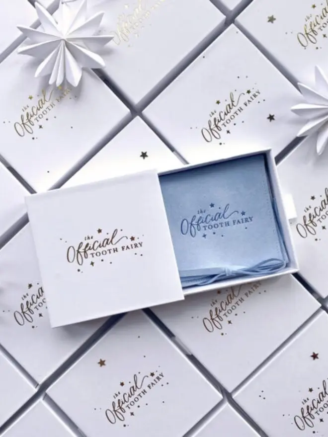 An open white slide-out box holds a pale blue suede pouch embossed with "The Official Tooth Fairy." It sits on rows of matching gold foil gift boxes with white paper stars.