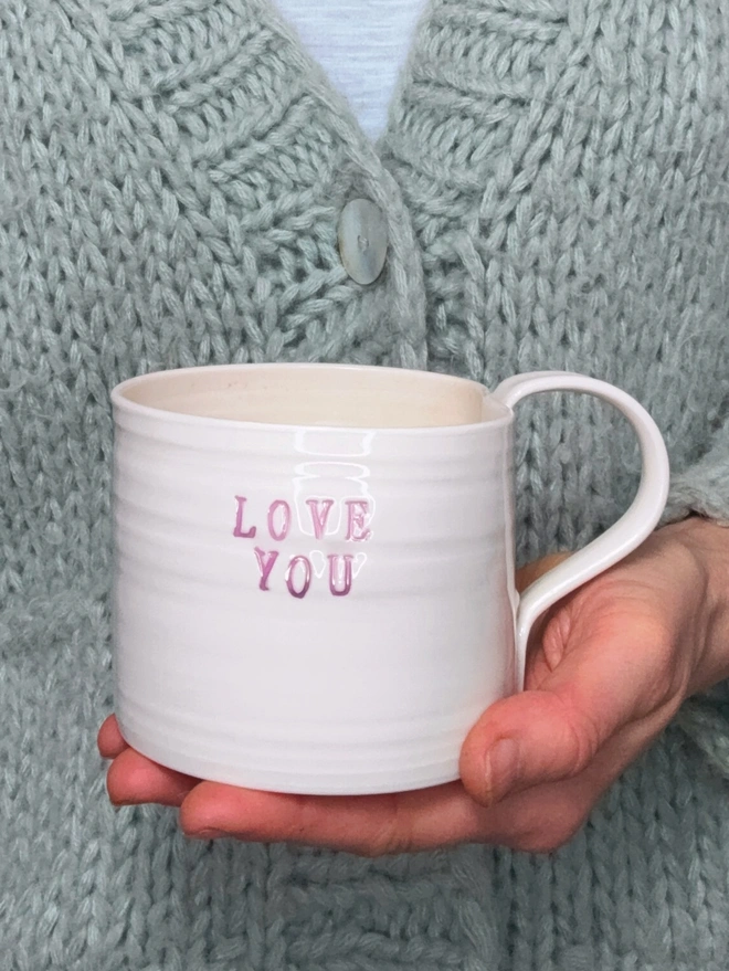 A hand thrown porcelain mug with the words LOVE YOU stamped into the surface in pink lustre. The mug is held in one hand against a sage coloured woollen cardigan