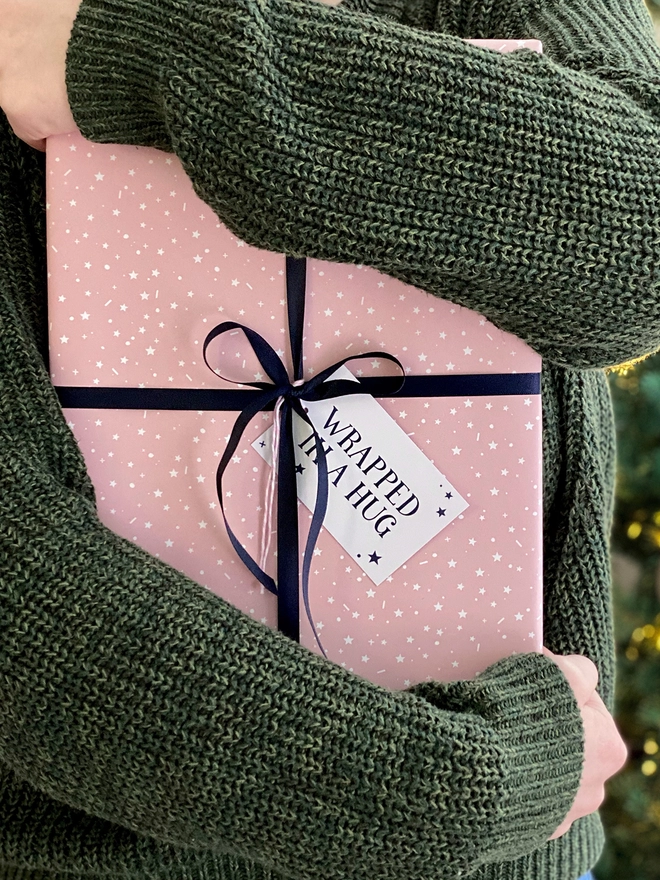 A gift wrapped in a gentle pink start design wrapping paper, tied with navy blue ribbon, with a gift tag that reads "Wrapped in a hug" is being held in front of a Christmas Tree.