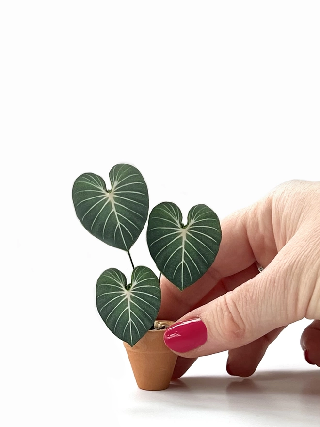A miniature replica Philodendron Gloriosum paper plant ornament in a terracotta pot held between 2 fingers against a white background