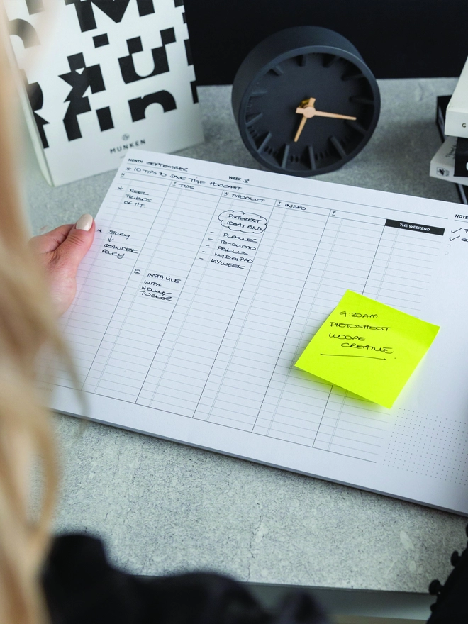 Lady holding a Hello Time Weekly Planning pad with a yellow post-it note on a desk with a clock