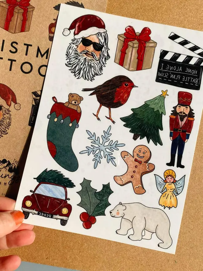 Christmas Temporary Tattoos, a hand holds a brown kraft paper booklet titled "CHRISTMAS TATTOOS" next to a sheet of festive holiday temporary tattoos.