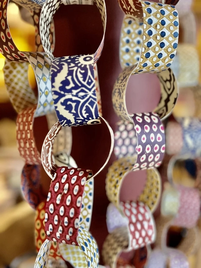 Colourful Paperchains (Set Of 60). Close-up of hanging paper chain garlands made from patterned links in navy, ochre, rust and red. The block-print-style designs make a colourful handmade decoration for festive or everyday styling.