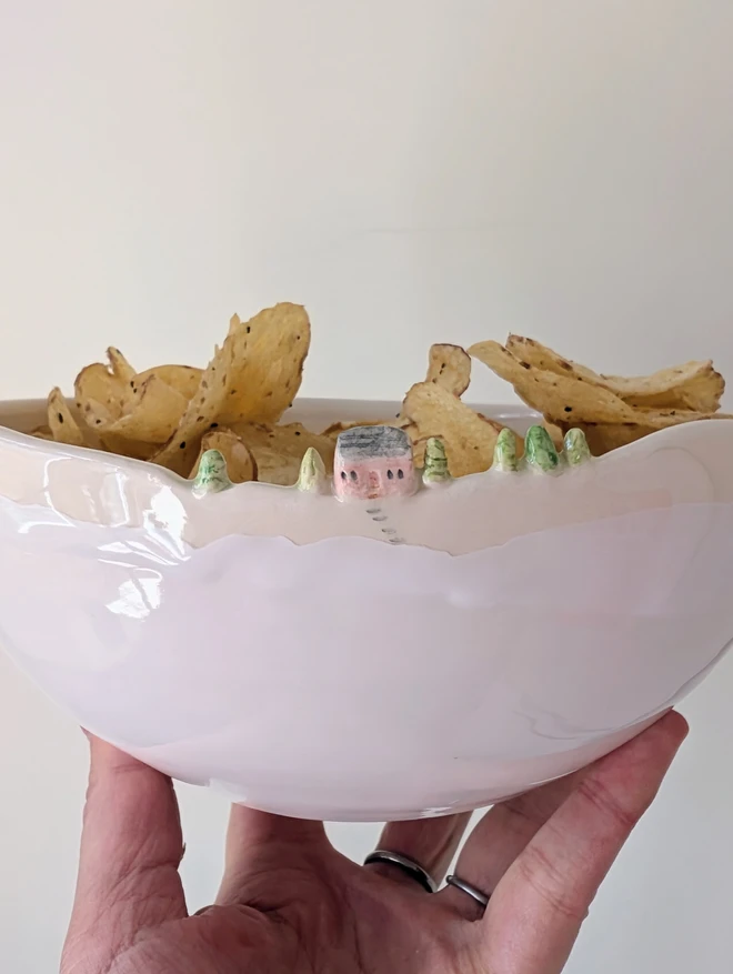 Handmade ceramic white bowl with a tiny pink house and tree details on the rim and white glaze held aloft in a hand