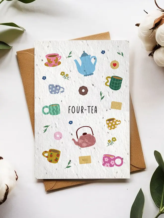 A greeting card titled “FOUR-TEA” featuring colorful illustrations of teapots, mugs, flowers, and tea bags, placed on top of a brown envelope surrounded by cotton flowers and green leaves on a white background.