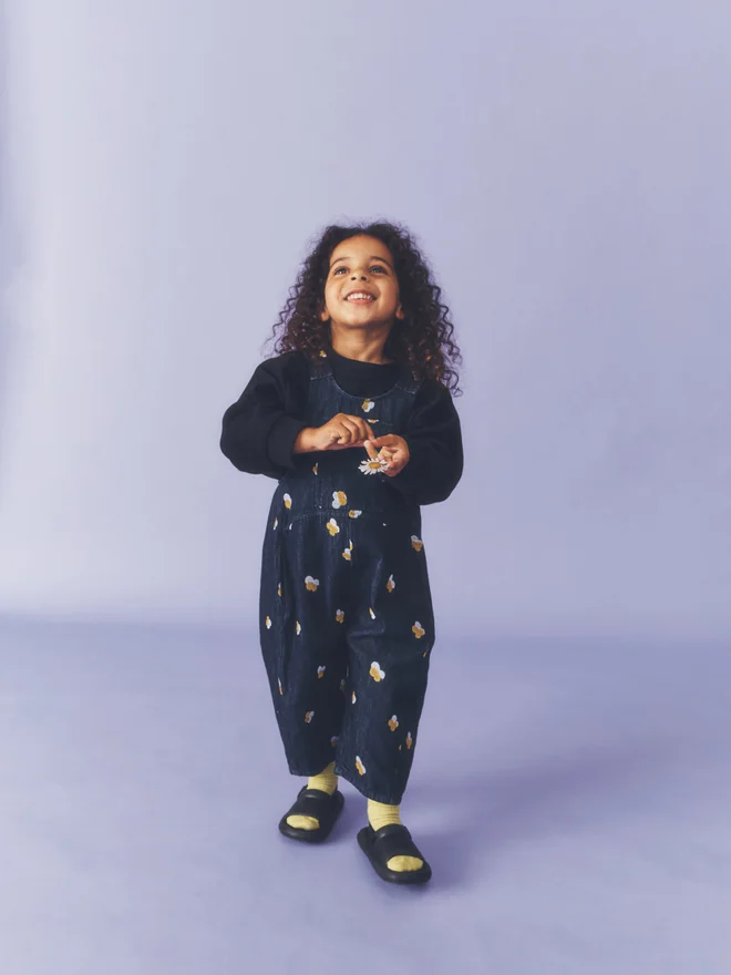 Close crop of a child in a rust ribbed beanie smiling against a lilac backdrop, wearing Studio Koter Kai dungarees in Denim Petal over a black sweatshirt.