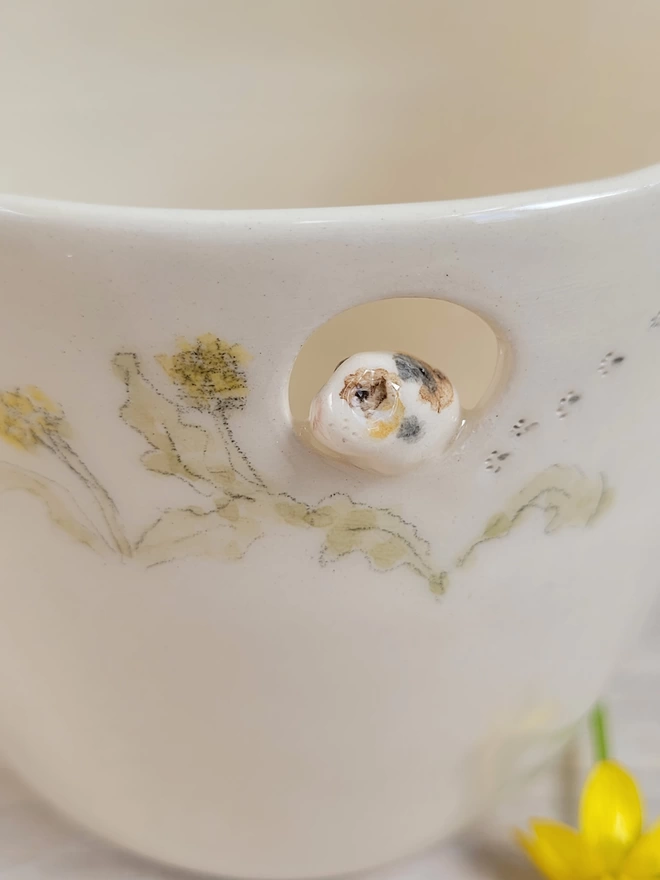 close up of a tri colour ceramic guinea pig sitting in a cut out circle in a handmade pottery cup with delicately painted dandelions and leaves