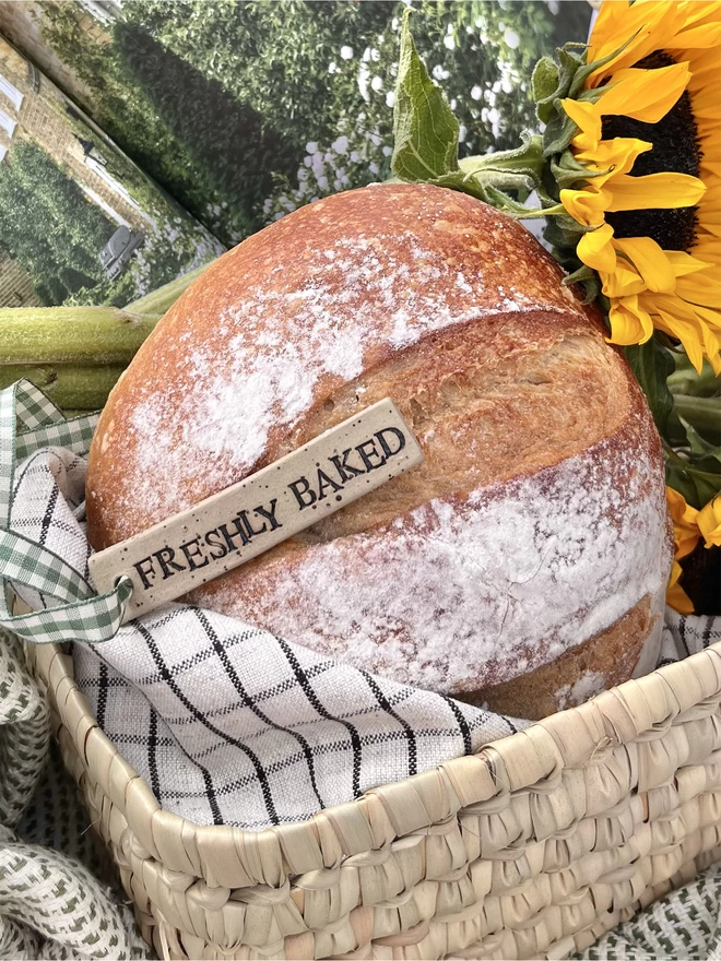 large round loaf of bread wrapped in a tea towel with a ceramic tag laid across it, featuring the word 'freshly baked'