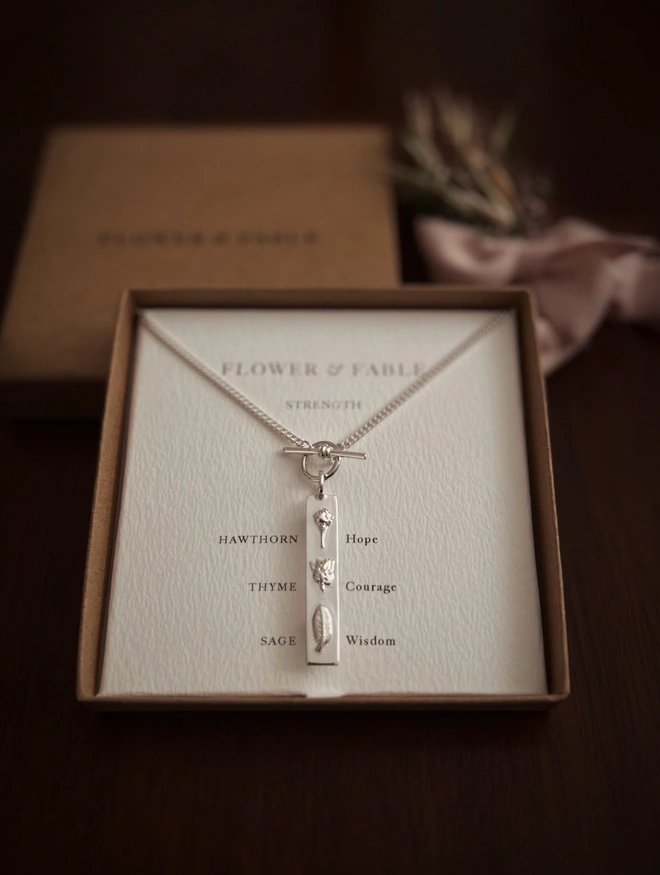 Strength bar pendant on a curb t bar necklace in sterling silver in a box with the flower meanings on a dark background
