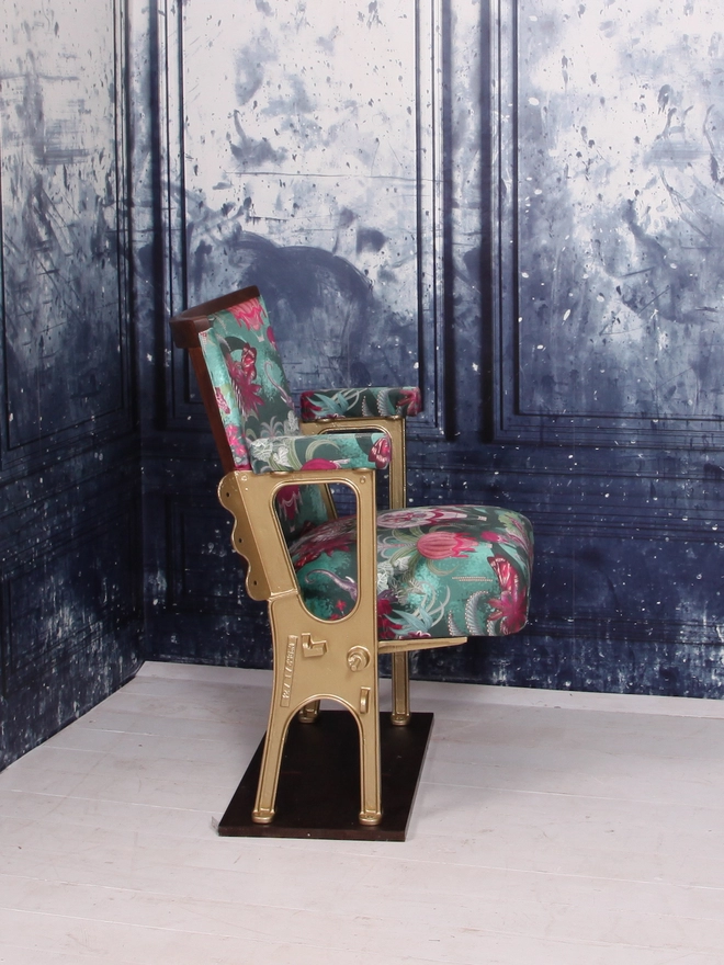 Side view of a single vintage cinema seat upholstered in a green and pink safari velvet, against a blue marbled wall.  The seat of the cinema seat is down