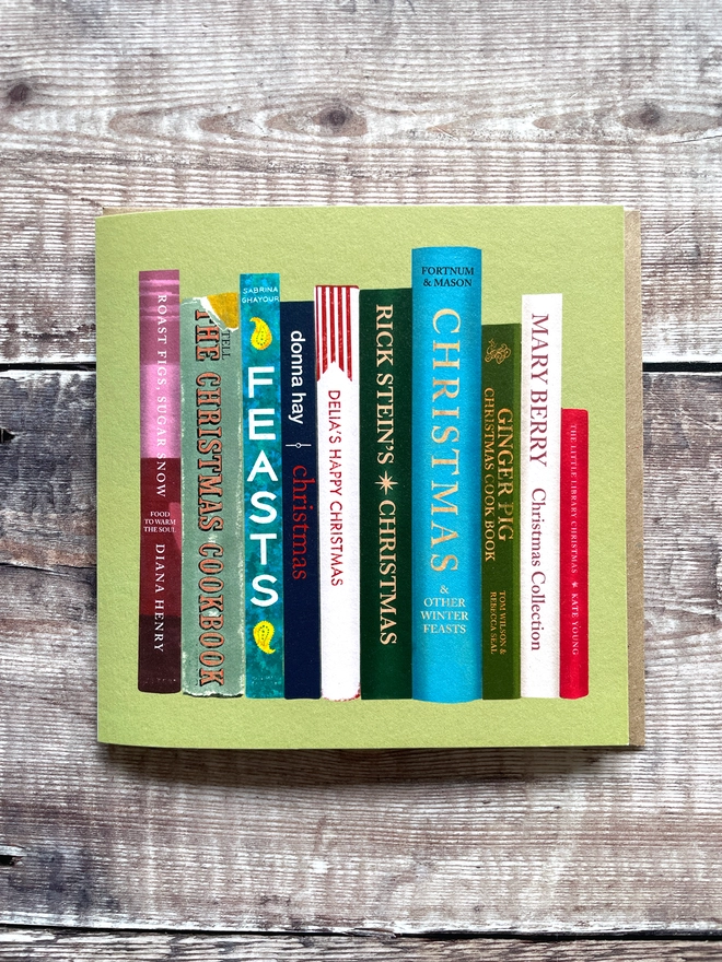 Green Christmas card illustrated with a stack of colourful cookbook spines, including titles styled after Nigella's Feasts, Delia's Happy Christmas, Rick Stein's Christmas, Mary Berry's Christmas Collection, and Fortnum & Mason Christmas, on a rustic wood background.