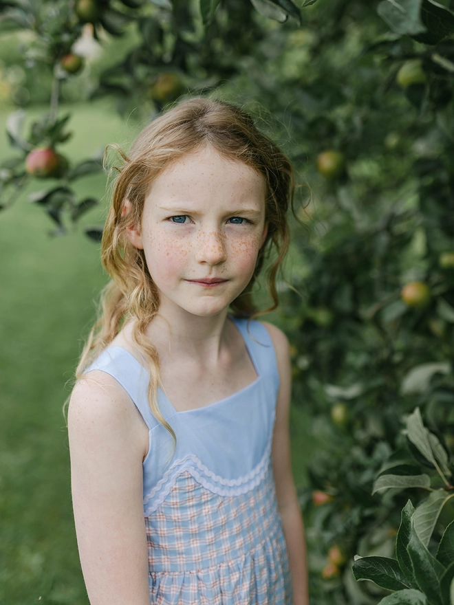 A girl standing near an apple tree wears a blue checked sundress with rikrak detail. 