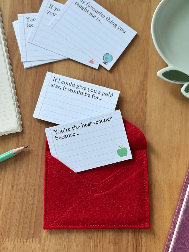 A handmade red felt apple-shaped envelope lays upside down. The heirloom apple envelope is surrounded by prompt cards that can be completed by a child, displayed on a wooden desk. It's a thoughtful gift for teachers from children.