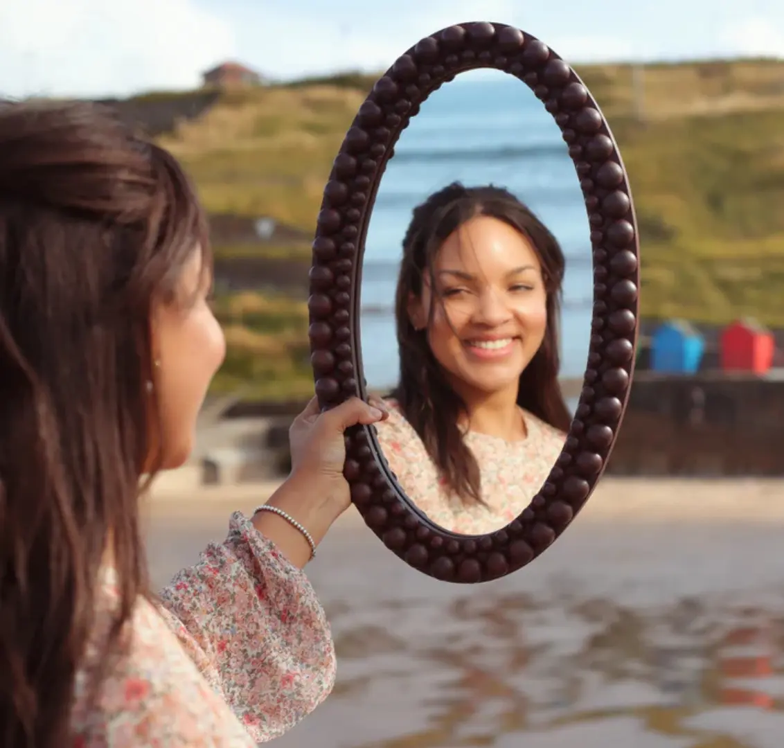 Woman holding an oval bobbin mirror on a beach with her face reflected in the mirror.