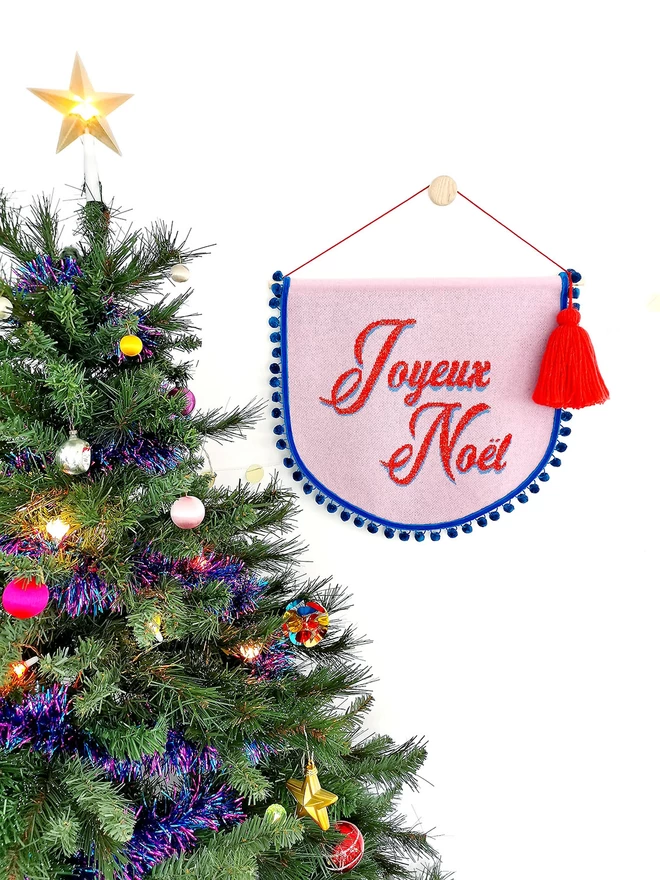 A pink knitted scallop shaped wall hanging with teal pom pom trim hangs on a wall next to a colourful Christmas tree hung with retro glass baubles and a lit up star on top.. The words ‘Joyeux Noel’ are written across the banner in red retro writing and an oversized red tassel hangs from one side of the wooden doweling.