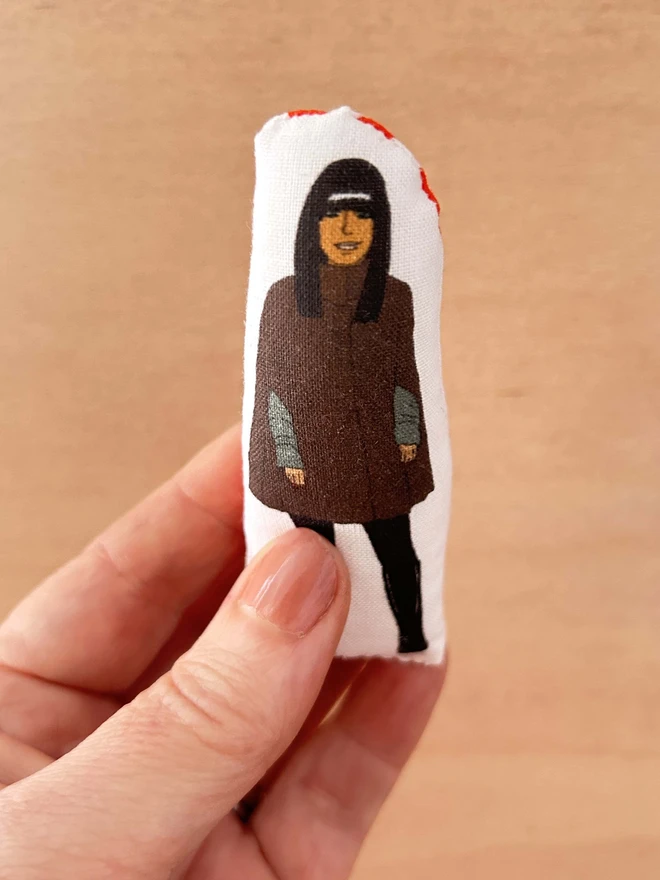 Mini fabric doll featuring an illustration of Claudia Winkleman in her Traitors cape, held up against a plain backdrop.