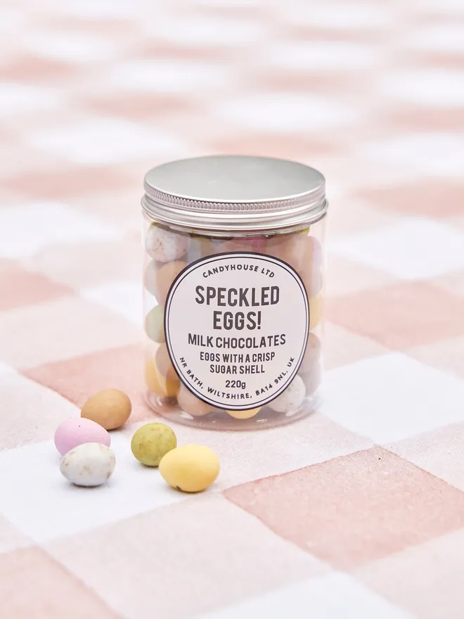 A clear jar of milk chocolate speckled eggs with a crisp sugar shell on a pink and white checkered tablecloth with a few speckled eggs on the side 