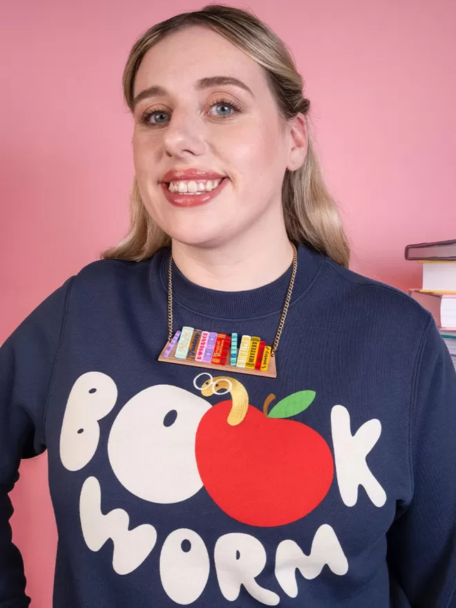 Model is wearing the tatty devine world book day bookshelf necklace. She is holding some books and wearing the necklace over navy jumper with a bookworm slogan.