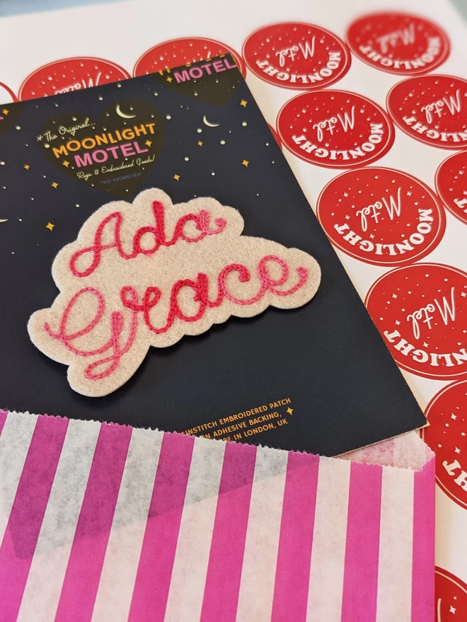 Ivory felt patch with Pink lemonade coloured gradient embroidery thread stitched to read the name 'Ada Grace', presented on a blue backing card and surrounded by stripey packaging and Moonlight Motel stickers ready for shipping