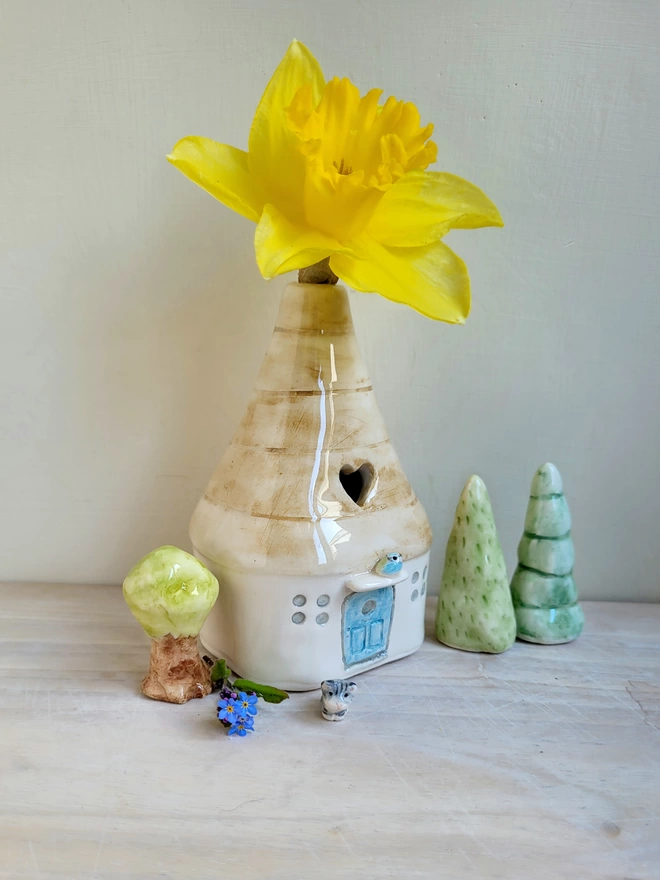 pottery vase in the form of a house with a bluetit perched above the door and green ceramic tree models each side of the vase and there is a daffodil in the vase 