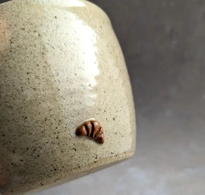 Biscuit coloured croissant coffee cup with a subtle croissant design, placed against a grey marble background.