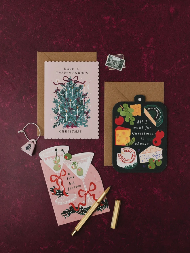 Tree-mendous Christmas card on a burgundy background with the Cheeseboard and Tini Bit Festive Christmas cards and a gold pen