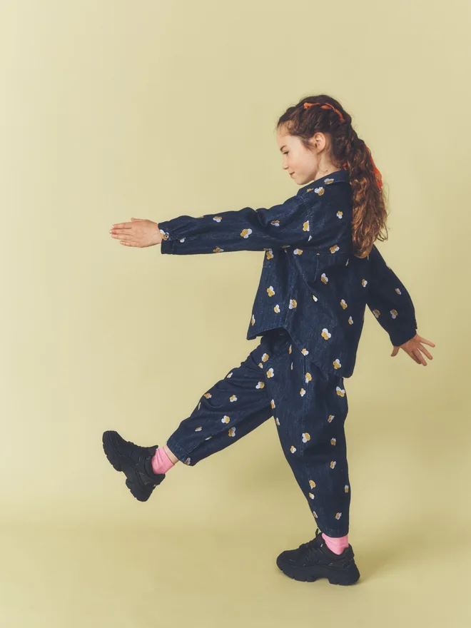 Side view of a child mid-stride with arms outstretched against a pale yellow backdrop, wearing a matching Studio Koter embroidered dark denim overshirt and trousers.