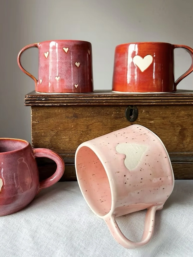 Heart Mug Strawberry Pink, a pink mug on a white surface in front of more red heart mugs, on a small wooden cabinet. 