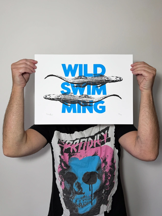 A humorous limited edition A3 screen printed poster. Featuring black alligators over blue 'wild swimming' typography. Scale shot of poster.