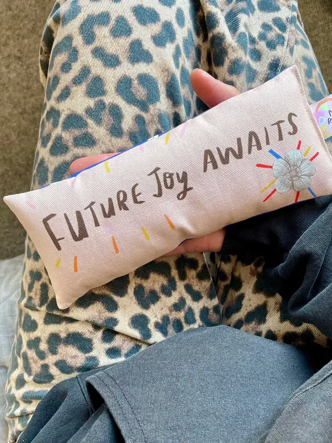 Model holds peach coloured lavender eyebag which reads 'future joy awaits'