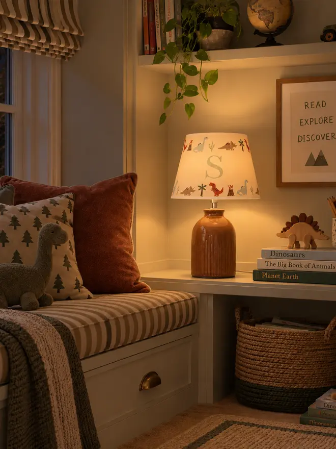 Cosy children’s reading nook at night with a personalised dinosaur lampshade featuring the initial S, styled with dinosaur books, toys and cushions.