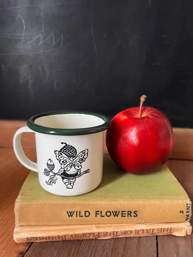 a desk setting with stacked books, and a miniature enamel mug cream in colour with a bottle green rim featuring an illustration of a small acorn and pixie on a branch