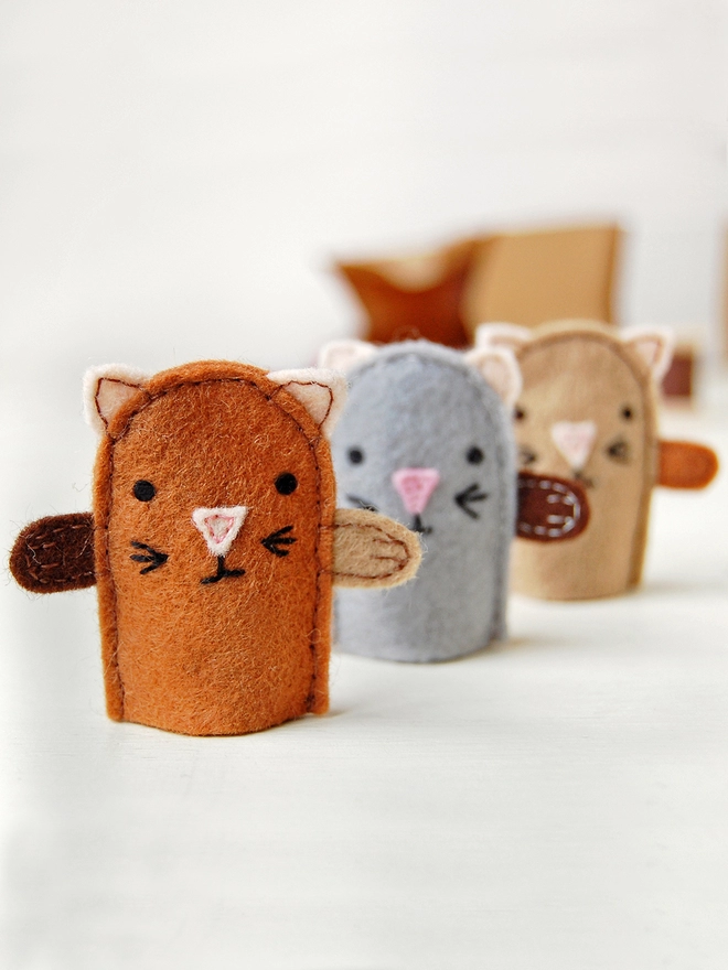 Three felt kitten finger puppets stand on a white desk in front of a craft kit to make them.
