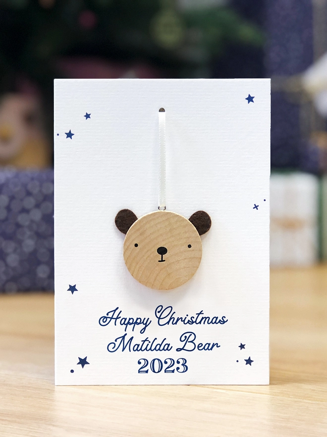 A personalised Christmas greetings card with a small wooden bear decoration attached is being held in front of a Christmas tree.