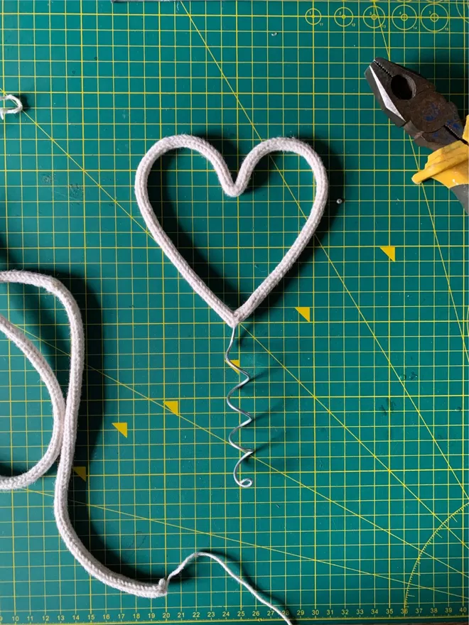 A white string wrapped wire heart shaped Christmas tree topper on a green cutting mat with tools