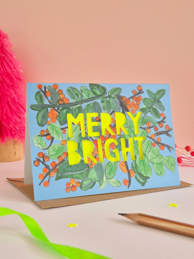 Merry Bright Christmas Card with Orange Berry Illustration, paper cut text with neon yellow slogan that says Merry Bright 