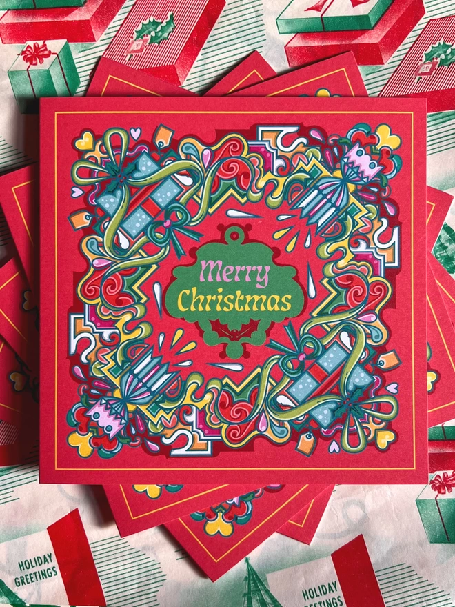 Three square cards displayed on top of each other, sitting on Christmas wrapping paper. An abstract Christmas card design, with Merry Christmas in the centre, surrounded by a multi-coloured design including crackers and presents, on a red background.