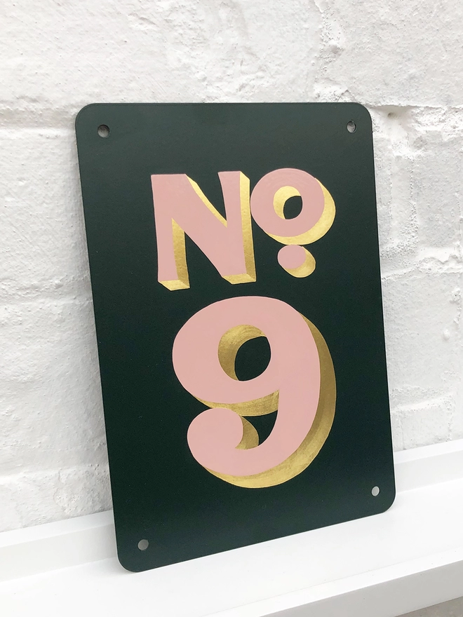 Hand painted house number No. 9 in pale pink and gold leaf on anthracite grey metal plaque, against a white brick wall. 