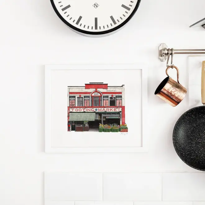 Watercolour illustration of Tooting Market, framed in white and hanging below a clock on a white kitchen wall
