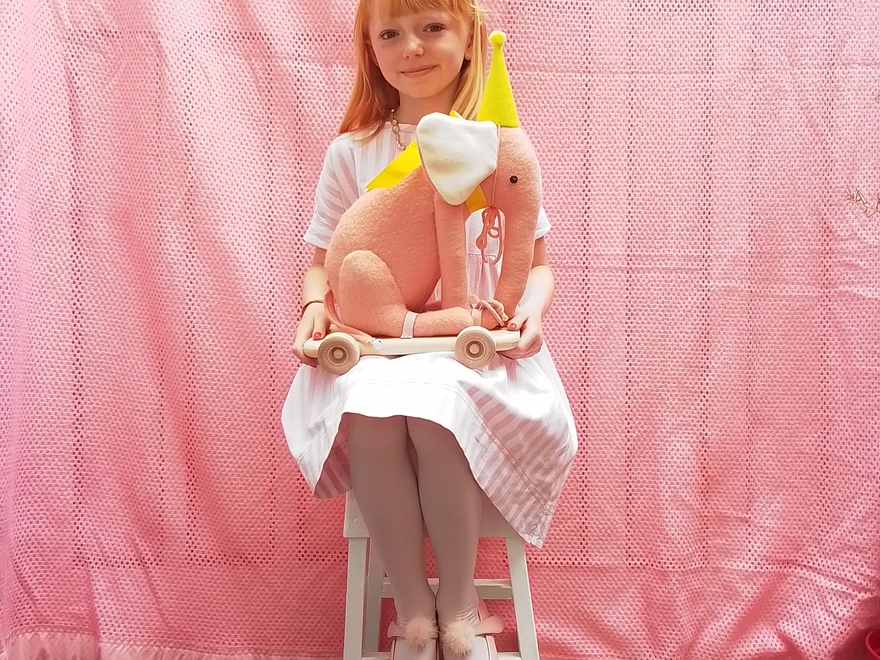 A girl sat on a stool, holding a pink pull-along elephant toy, wearing a yellow party hat