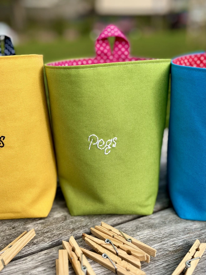 Three brightly coloured hanging cotton peg bags with 'Pegs' embroidered on the front, standing on a wooden bench with clothes pegs scattered about, in a garden.