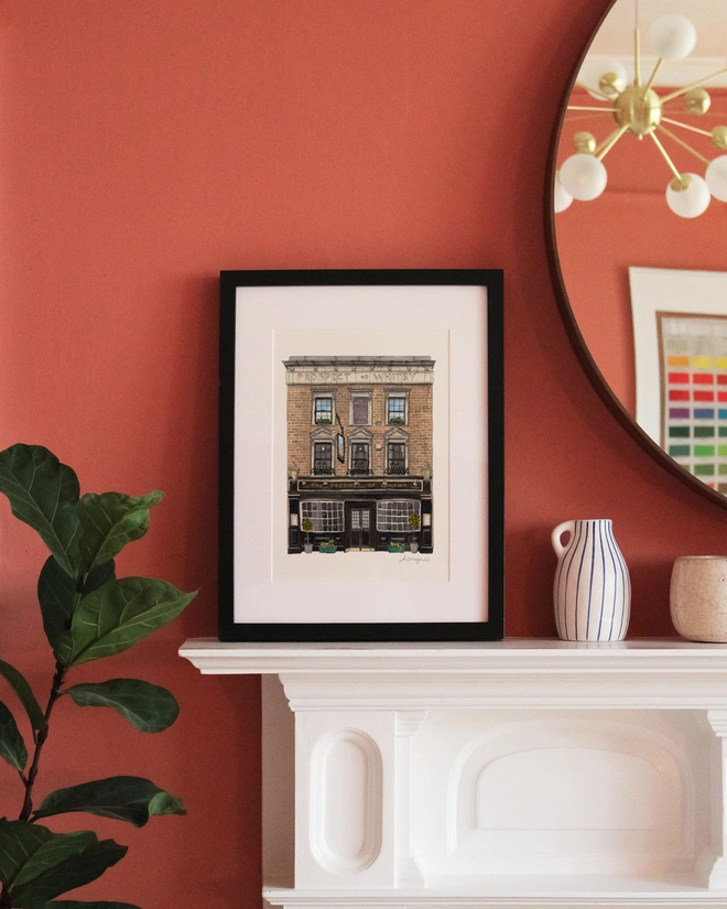 A framed print featuring an illustration of The Prospect of Whitby pub displayed on a white mantelpiece against a terracotta-colored wall.