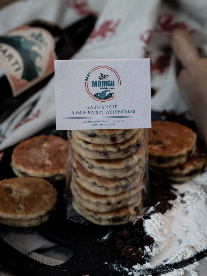 Barti Rum and Raisin Welsh Cakes and loose welsh cakes on black griddle surrounded by loose ingredients with a welsh dragon and beige table cloth in the background