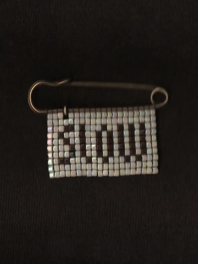 'lingua i' beaded 'slow' pin. a beaded pin badge with the word slow in brown lettering on a pearl background.