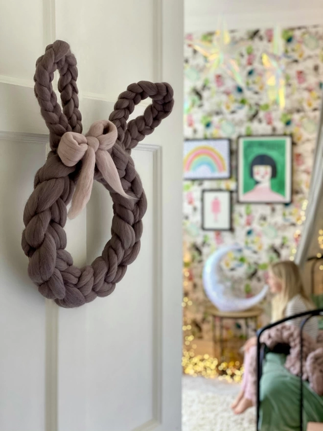 A soft grey woolly rabbit wreath is hung on the door of a little girl's room, you can see her colourful wallpaper, prints and soft fairy lights through the door
