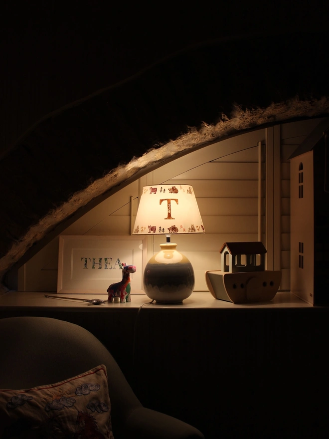 Children’s bedside table lamp with a personalised initial “T” lampshade, softly glowing in a cosy nursery setting. The handmade lampshade features small illustrated animal motifs around the rim, creating warm ambient lighting beside wooden toys and a dollhouse under a sloped ceiling.