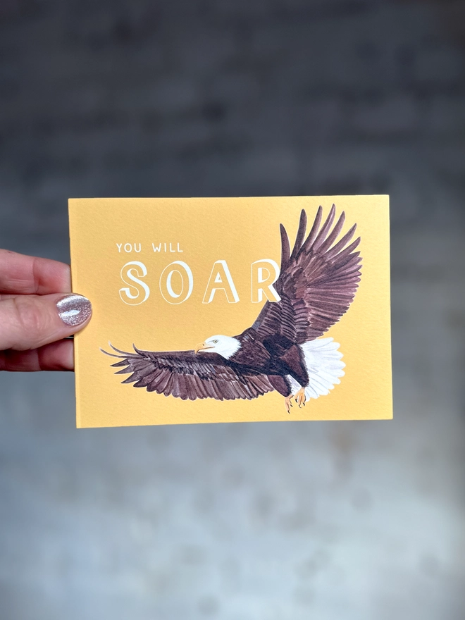a bright yellow greetings card featuring an illustration of a bald eagle with the text “you will soar”